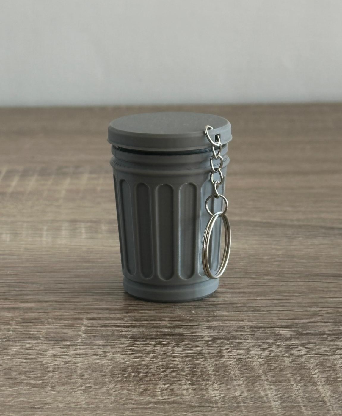 Porta-chaves em forma de caixote do lixo cinza com tampa e argola metálica, sobre superfície de madeira.