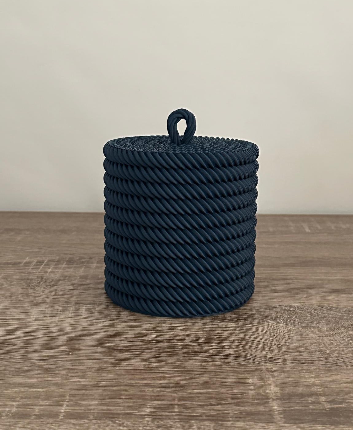 Recipiente cilíndrico de corda preta com tampa e pega em laço sobre mesa de madeira.
