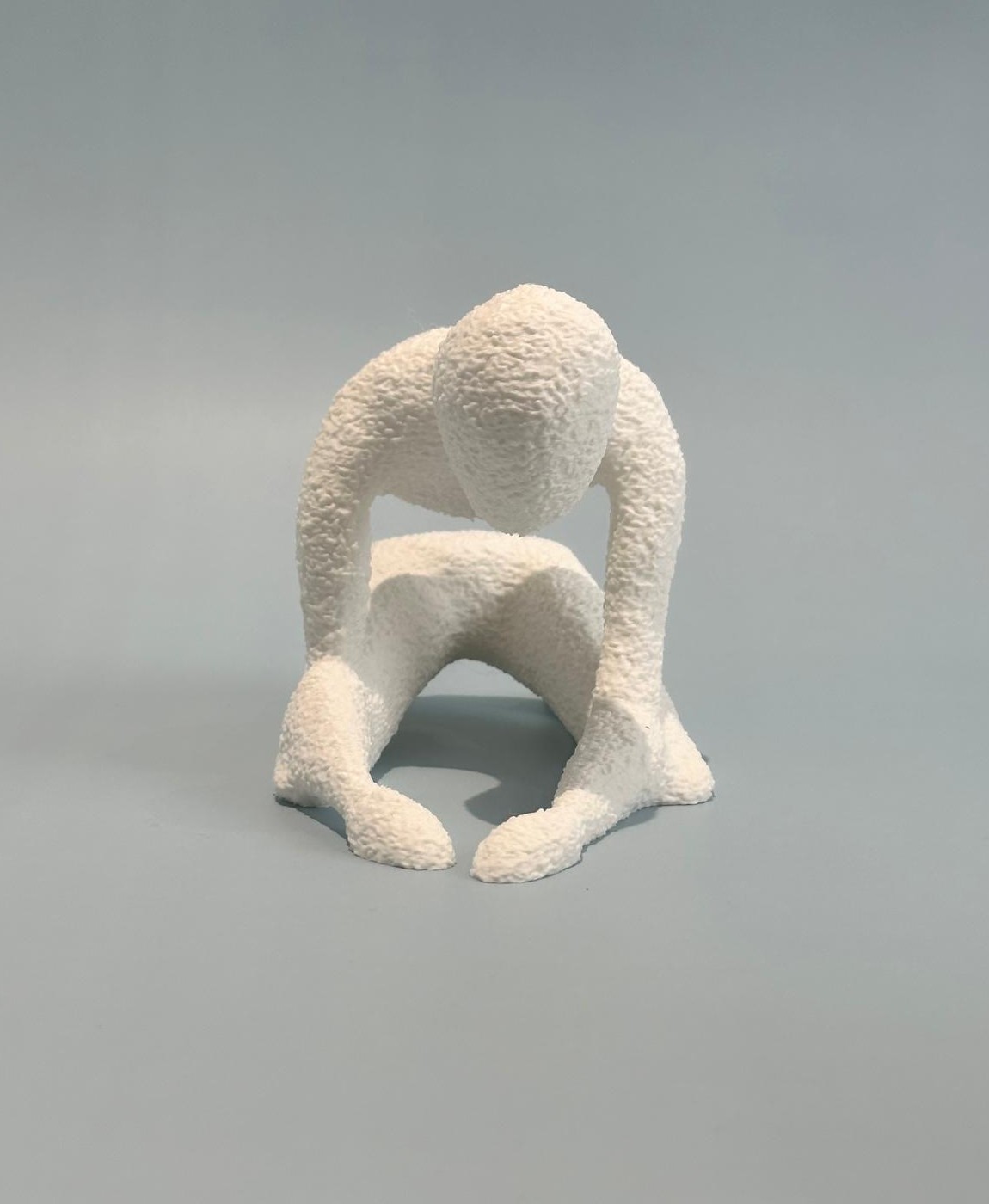 Escultura branca rugosa de figura humanoide agachada sobre fundo cinzento