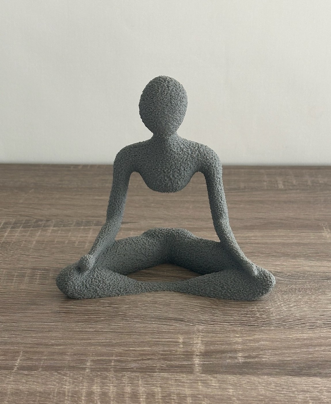Estatueta cinzenta rugosa em forma de figura humana sentada, sobre mesa de madeira clara