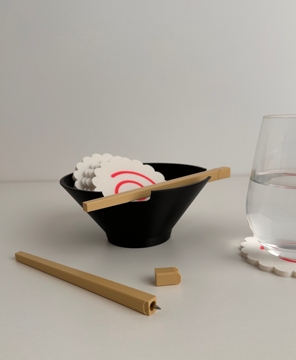 Taça preta com bases de copos brancas com padrão de flores e círculo vermelho, palitos de madeira cor bege, vidro de água com base de copo no fundo cinzento