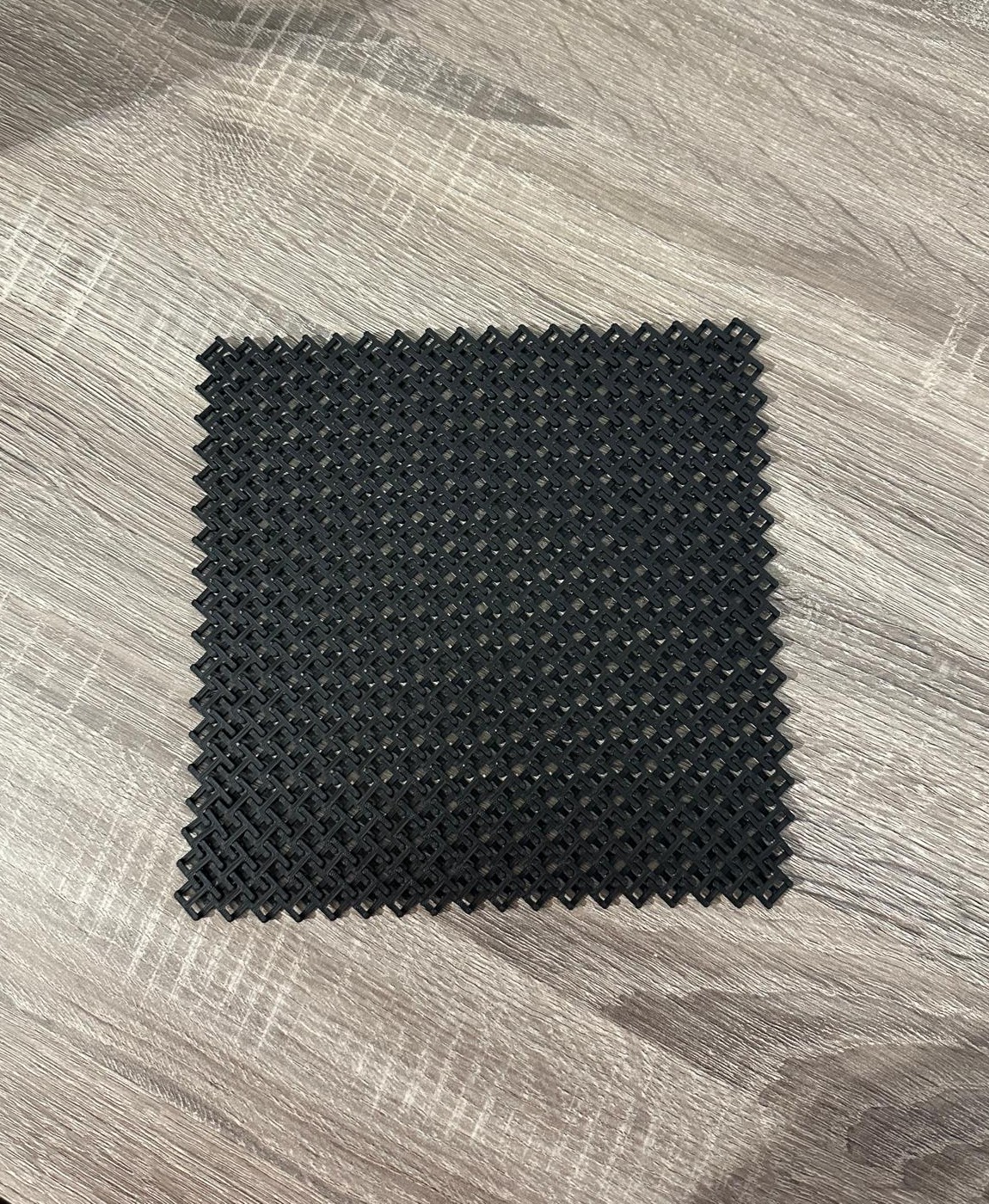 Peça quadrada de plástico preto com padrão perfurado sobre madeira clara