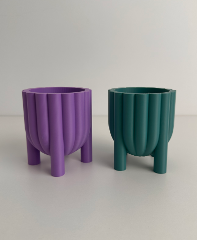 Dois vasos pequenos, um roxo e outro verde-azulado, com textura vertical e três pés, sobre superfície branca