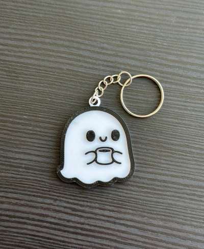 Chaveiro fantasma branco com xícara, anel metálico dourado, sobre superfície cinza escuro