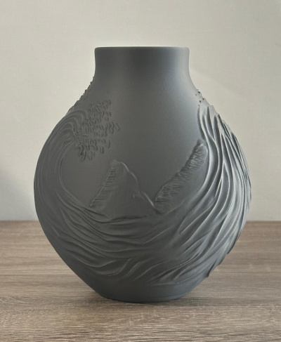 Vaso cinzento fosco com relevo texturizado de ondas e montanha sobre mesa de madeira clara