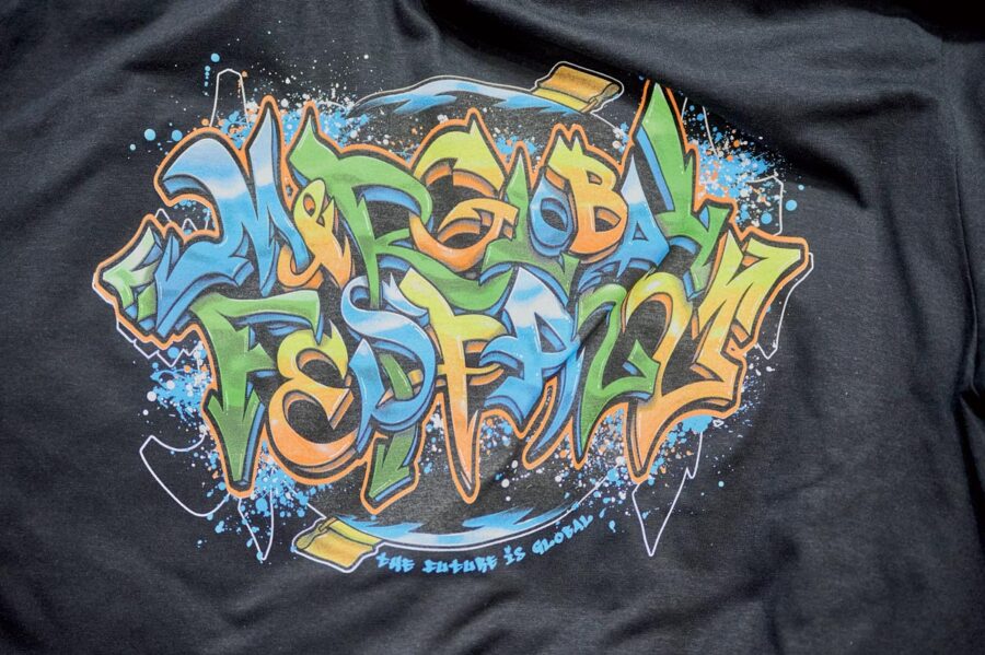 T-shirt preta com grafismo colorido de graffiti com texto 'MANGABA FESTIVAL' e 'SAFE BEHIND IS NORMAL'.