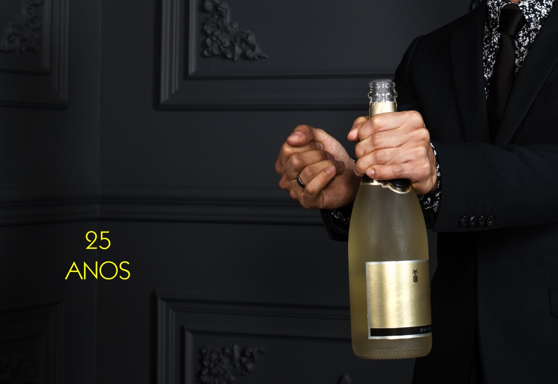 Pessoa de fato segurando uma garrafa de champanhe, fundo cinzento com texto 25 ANOS