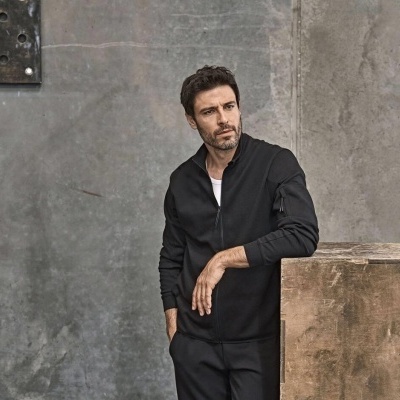 Homem com jaqueta preta com fecho de correr e bolso no braço, apoiado em caixa de madeira