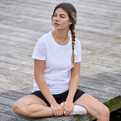 Mulher sentada de pernas cruzadas em madeira com t-shirt branca, calções pretos e ténis brancos.
