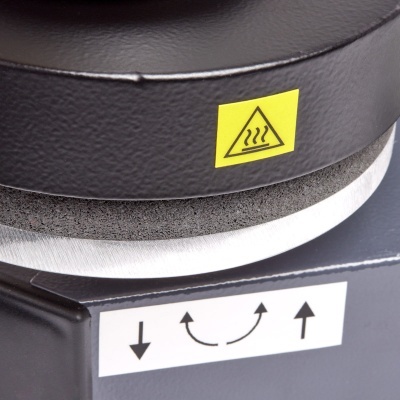 Detalhe de máquina preta com etiqueta amarela de aviso de calor e etiqueta branca com setas.