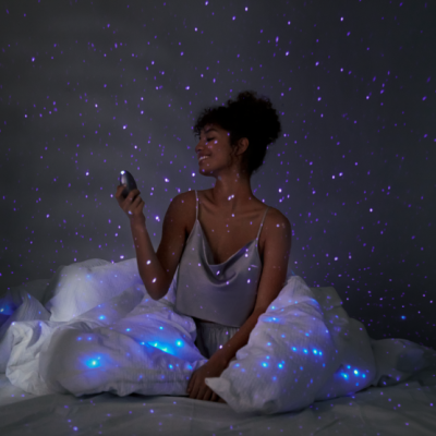 Mulher sentada na cama com dispositivo luminoso que projeta luzes azuis e roxas