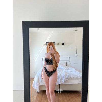 Mulher em lingerie preta rendada refletida num espelho num quarto com cama e parede branca.