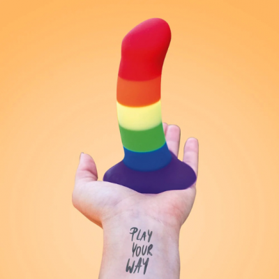 Dildo colorido em tons de arco-íris na mão com tatuagem 'PLAY YOUR WAY' em fundo laranja