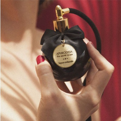 Frasco de perfume preto com pulverizador dourado e laço, mão de mulher com unhas vermelhas.