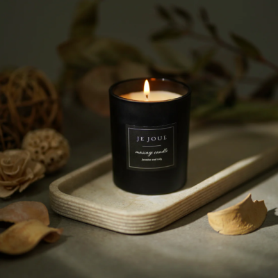 Vela perfumada acesa em recipiente preto com rótulo 'JE JOUE massage candle Jasmine and lily' numa bandeja de madeira clara.