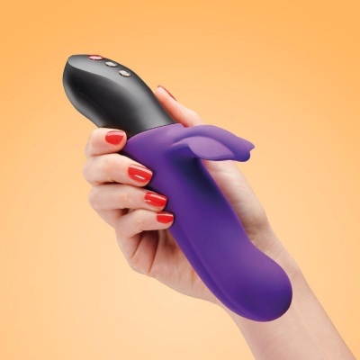 Vibrador roxo com parte superior preta e botões, segurado por uma mão com unhas vermelhas