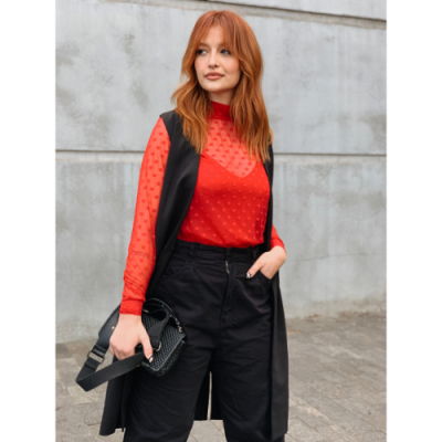 Mulher ruiva com blusa vermelha de pontos e colete preto num fundo de parede cinza