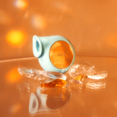 Dispositivo azul claro com frente laranja metálica LELO e cristais translúcidos sobre superfície refletiva laranja