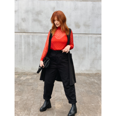 Mulher com blusa vermelha, calças e colete pretos, botas e mala preta, em fundo cinzento