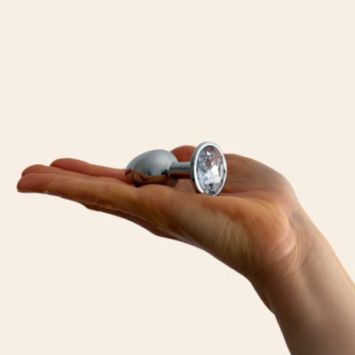 Plugue anal metálico prateado com pedra transparente na ponta, segurado por mão.