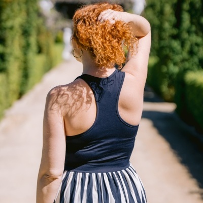 Pessoa de costas com vestido azul escuro e saia listrada, cabelo ruivo, ao ar livre com vegetação