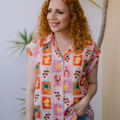 Blusa feminina colorida com padrão de frutas e lagostas em fundo branco e parede branca com planta
