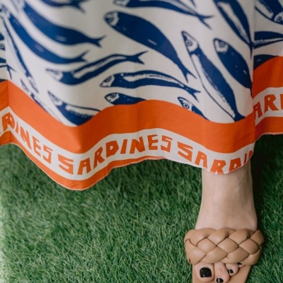 Vestido branco com padrão de sardinhas azuis e faixa laranja com texto e pé com sandália trançada bege na relva