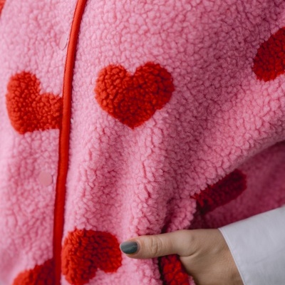 Casaco peludo rosa com corações vermelhos e rebordo vermelho, mão segurando o casaco com unhas cinza.