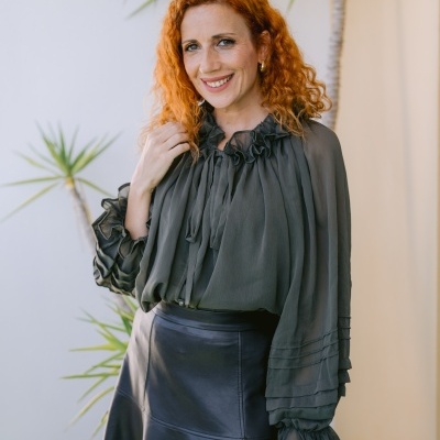 Mulher com blusa verde escuro transparente com folhos e saia preta de couro, em fundo com plantas