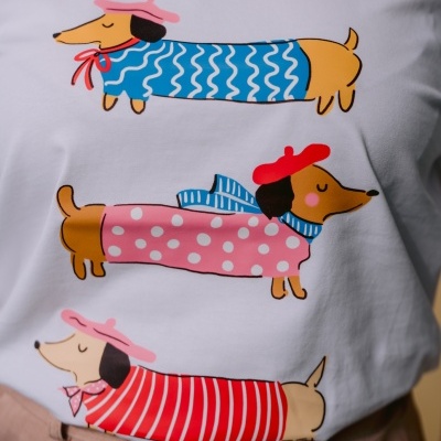 T-shirt branca com ilustrações de três cães tipo dachshund em camisolas coloridas.