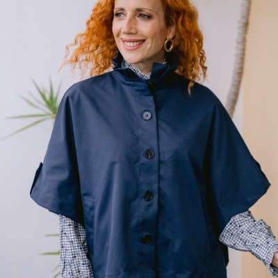 Mulher com capa azul, camisa xadrez e jeans, de cabelo ruivo encaracolado.