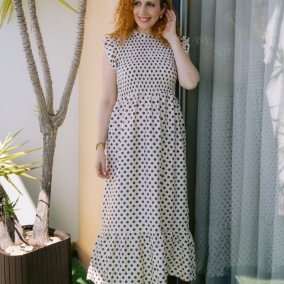 Vestido branco com bolinhas pretas usado por mulher ruiva em exterior junto a planta e janela.