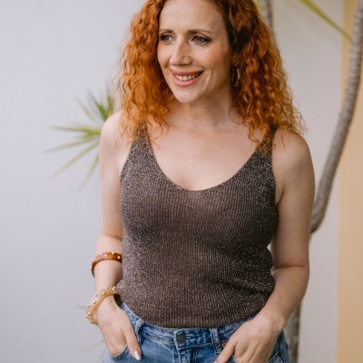 Mulher de cabelo ruivo cacheado a usar top castanho tricotado com brilho e jeans azuis de cintura alta com pulseiras castanhas.