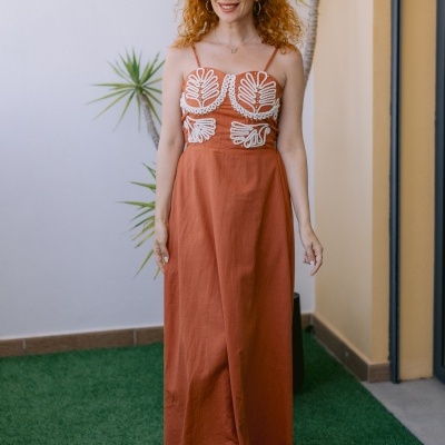 Vestido longo terracota com bordados brancos e alças finas, vestido por mulher com cabelo ruivo confiante em ambiente exterior.