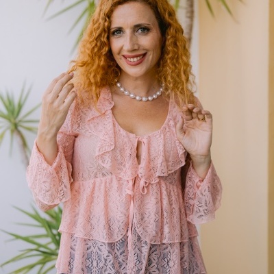 Mulher com blusa de renda rosa clara e colar de pérolas em ambiente com plantas