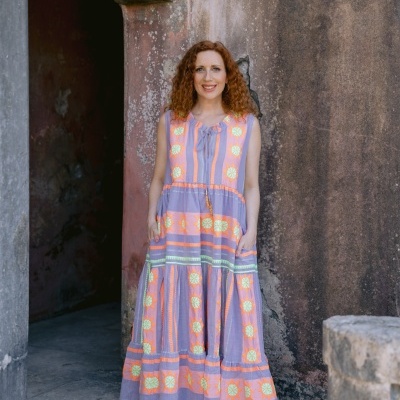 Vestido comprido lilás com padrões florais e cordão, mulher ruiva a sorrir em frente a parede envelhecida de pedra