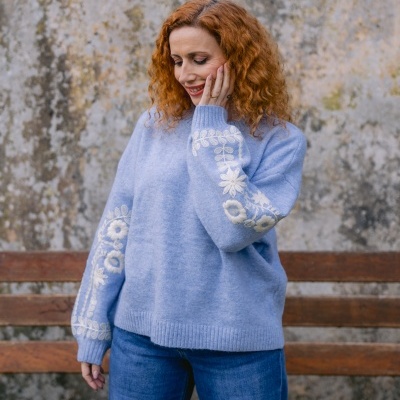 Suéter azul claro com bordado floral branco nas mangas, mulher com cabelo ruivo, banco de madeira e parede envelhecida ao fundo