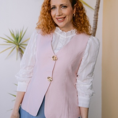 Colete rosa claro com botões dourados florais e blusa branca rendada, saia azul clara, planta verde no fundo