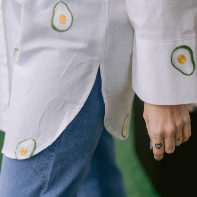 Camisa branca com bordados de abacate e calças de ganga azuis, braço direito com anel e tatuagem de coração.