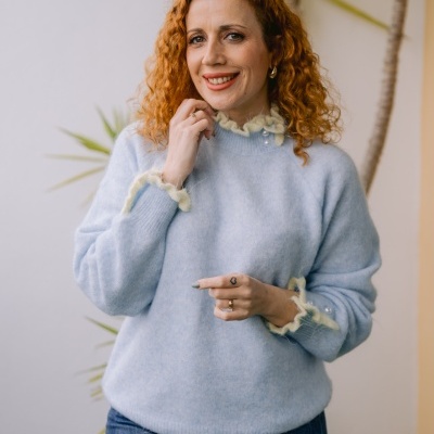 Camisola azul clara com detalhes creme e botões, mulher com jeans em fundo neutro com planta.