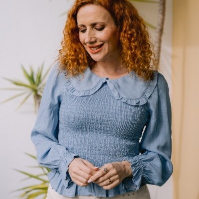 Mulher com blusa azul clara de manga comprida e calças bege claras com tachas