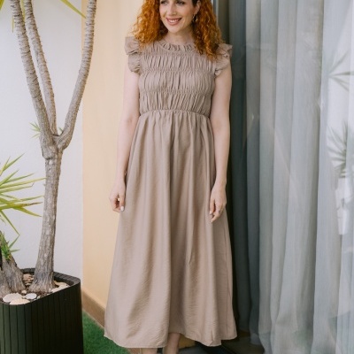 Mulher com vestido castanho claro e sandálias bege em ambiente exterior junto a plantas e vidro com cortinas.