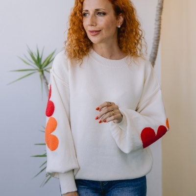 Mulher com camisola branca de malha com flores vermelhas e laranjas nas mangas a usar jeans azuis.