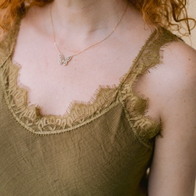 Top verde-oliva com renda na borda, colar dourado com asas e brincos de argola dourados em mulher com cabelo ruivo.