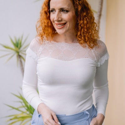 Mulher com blusa branca rendada e calças azuis claras junto a plantas.