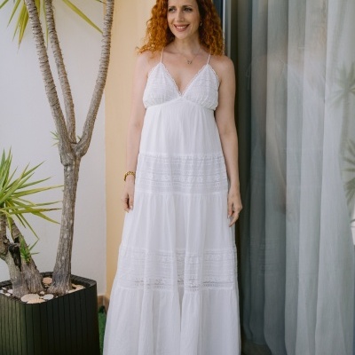 Mulher com vestido longo branco de tecido leve com detalhes em renda, ao lado de planta e janela com cortinas.