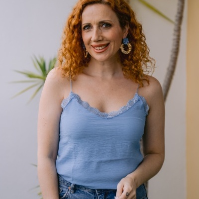 Mulher com blusa azul clara de renda e calças de ganga, com brincos dourados e flor azul, em ambiente com plantas