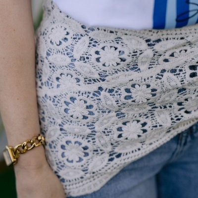 Pano de renda branco floral sobre calças de ganga azuis e pulseira dourada no braço.