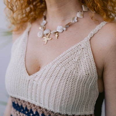 Pessoa a usar vestido com parte superior em crochet bege, castanho e azul, e colar com pingentes dourados e pérola