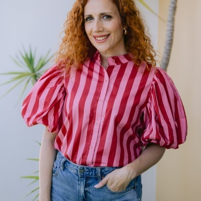 Blusa rosa e vermelha às riscas com mangas bufantes e calças de ganga azuis em ambiente com planta.